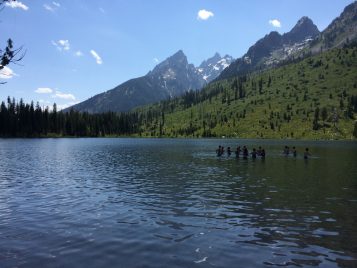 A color photograph of students in shadow standing in a lake. A sweeping view of sky and several peaked mountains rise out of the landscape behind them.